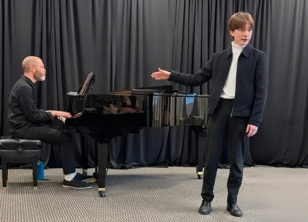 Oliver Kennedy performing beside a pianist at a grand piano, against a dark curtain.